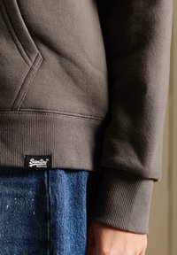 Sudadera con capucha hecha de una suave tela gris, con puños de canalé y una pequeña etiqueta negra de Superdry cerca del dobladillo. Lleva puesta encima de unos jeans de mezclilla azules.