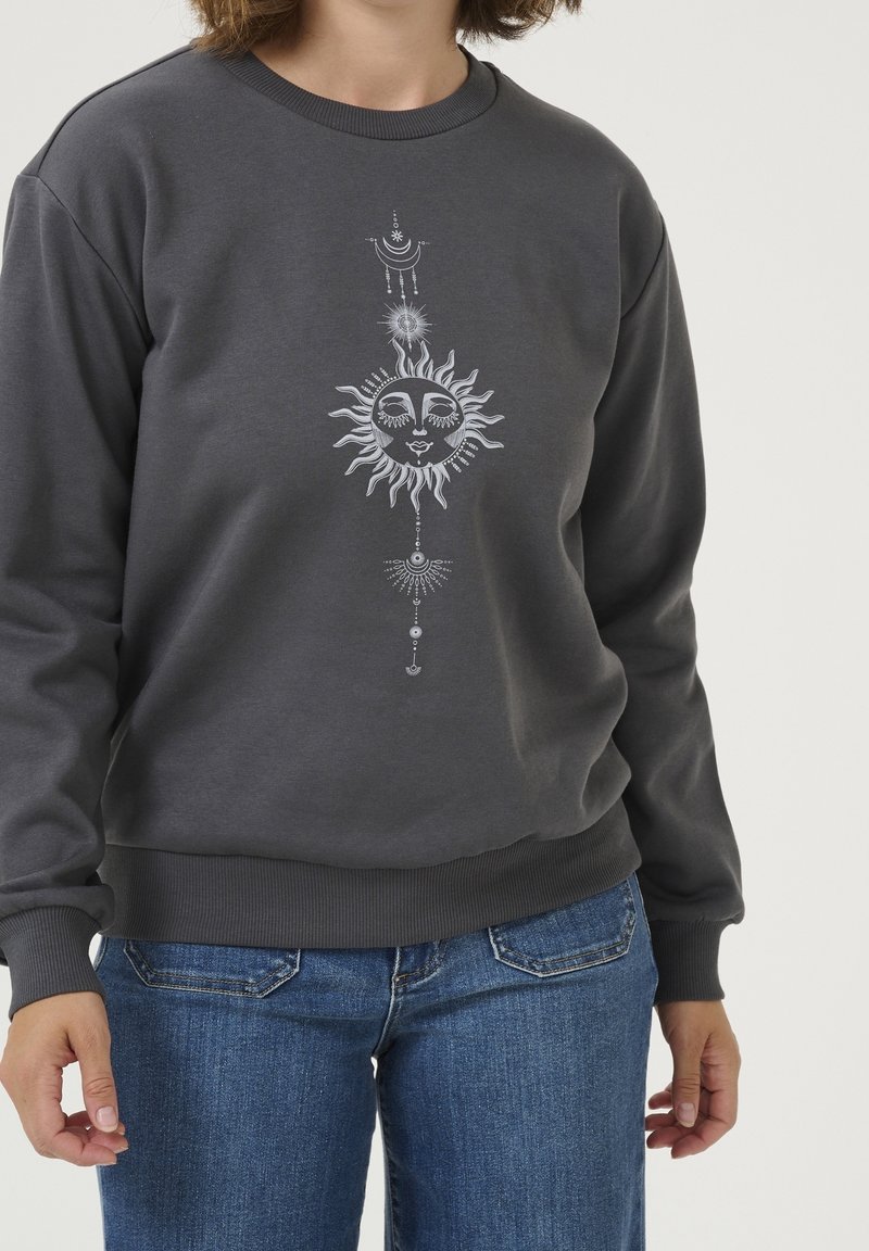 Femme portant un sweat-shirt gris foncé avec un motif blanc de soleil et de lune et un jean bleu, debout devant un fond clair uni.