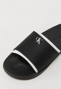 Musta liukusandaali, jossa on teksturoitu uritettu hihna, valkoinen korostusreunus ja metallinen logo. Pohja on valmistettu kestävästä materiaalista, jossa on kuvioitu pito.