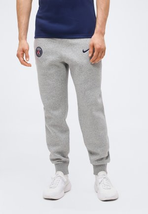 Mężczyzna noszący szare spodnie dresowe z logo Paris Saint-Germain i Nike, białe sneakersy oraz granatową koszulkę, stojący na białym tle.