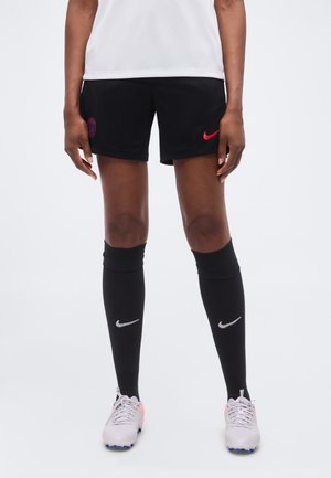 Person iført sorte Nike fodboldshorts med Paris Saint-Germain-logo, sorte Nike knæstrømper og hvide støvler, der står mod en hvid baggrund.
