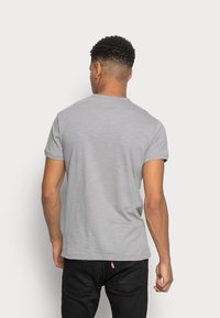 Man with short curly hair wearing a plain gray short-sleeve t-shirt and black jeans, standing facing away against a white background.