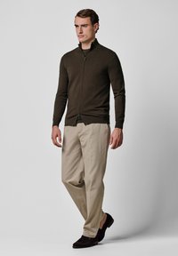 Ciemnooliwkowy sweter zapinany na zamek z kołnierzem stójką, noszony z beżowymi spodniami i ciemnymi butami, o dopasowanym kroju i ściągaczowych mankietach.