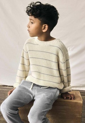 Pull en tricot crème avec des rayures grises, encolure ronde et coupe ample. Assorti à un pantalon de survêtement en polaire gris. Enfant assis sur une surface en bois.