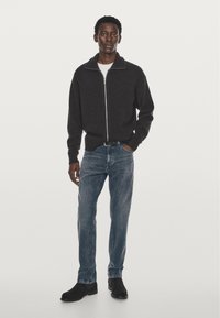 Donkergrijze zip-up sweater met een hoge kraag, gedragen over een wit shirt, gecombineerd met lichtblauwe jeans en zwarte suède laarzen.
