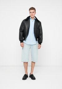 Fekete bőr bomberdzseki fekete gallérral, világoskék gombos ing, világoskék farmer rövidnadrág és fekete loafer cipő, egy sima háttér előtt állva.