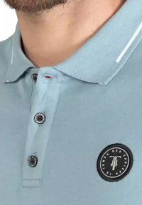 Lichtblauwe polo gemaakt van gestructureerde stof met een klassieke kraag, drie zwarte knopen en een cirkelvormig logo op de linkerkant.