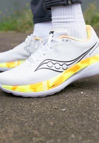 Tênis brancos de corrida com um cabedal em malha respirável, apresentam detalhes em preto e uma sola colorida que mistura tons de amarelo e laranja.