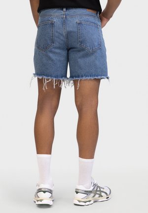 Person iført slidte blå denimshorts, hvide sokker og hvide sneakers, der står med ryggen mod kameraet på en ensfarvet baggrund.