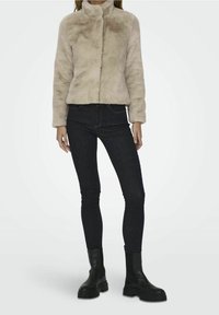 Beige fuskpälsjacka med hög krage, långa ärmar och dragkedja, kombinerad med mörk denim skinny jeans och svarta grova stövlar.