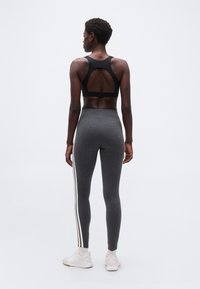 Γυναίκα που στέκεται με την πλάτη στην κάμερα, φορώντας μαύρο αθλητικό σουτιέν, σκούρο γκρι κολάν με λευκές ρίγες, λευκές κάλτσες και λευκά αθλητικά παπούτσια σε απλό φόντο.