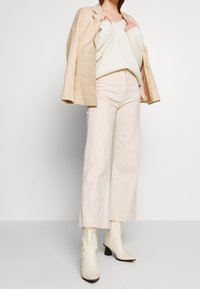 Femme portant un pantalon large beige, un pull en maille crème, un blazer à carreaux beige posé sur les épaules, et des bottines beige clair sur un fond uni.