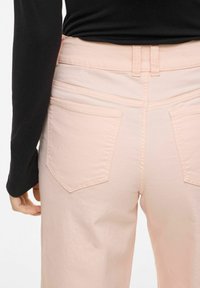 Gros plan sur une personne vêtue d'un pantalon rose pâle avec des poches arrière et un haut à manches longues noir, debout devant un fond uni.