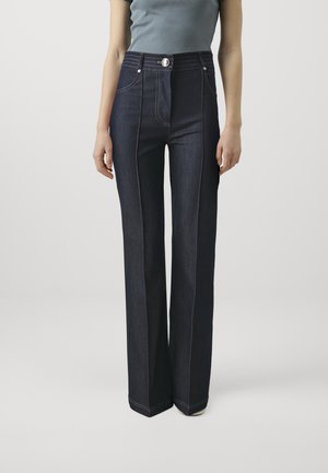 Kvinde iført højtaljede, mørkeblå jeans med vide ben, kontrastsyning og lukning med knapper, kombineret med en grå tætsiddende kortærmet top.