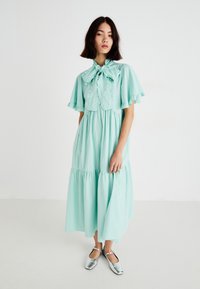 Vestido de algodón verde menta con mangas acampanadas y un cuello con lazo. Falda de capas en forma acampanada. Modelo lleva zapatos plateados metálicos. Fondo minimalista.