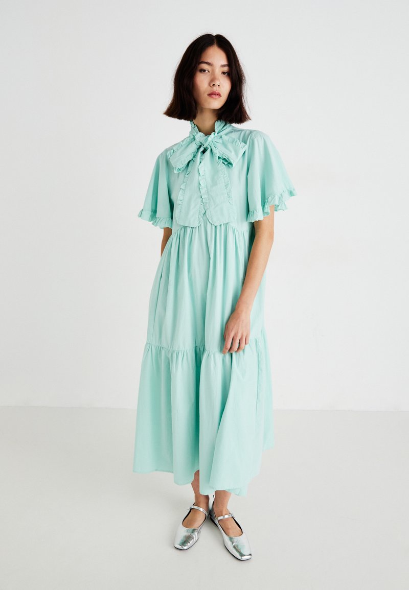 Vestido de algodón verde menta con mangas acampanadas y un cuello con lazo. Falda de capas en forma acampanada. Modelo lleva zapatos plateados metálicos. Fondo minimalista.