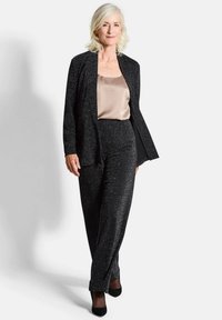 Femme aux cheveux courts blancs portant un costume noir brillant et un chemisier en satin beige, se tenant devant un fond clair.