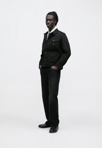 Čierna rifľová bunda s gombíkovými vreckami, v kombinácii s tmavými džínsami a čiernymi topánkami. Biela košeľa viditeľná pod bundou.