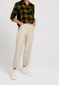 Chemise en flanelle à carreaux verts et jaunes avec boutons blancs, associée à un pantalon beige cintré et des baskets montantes blanches. Détail de poche simple.