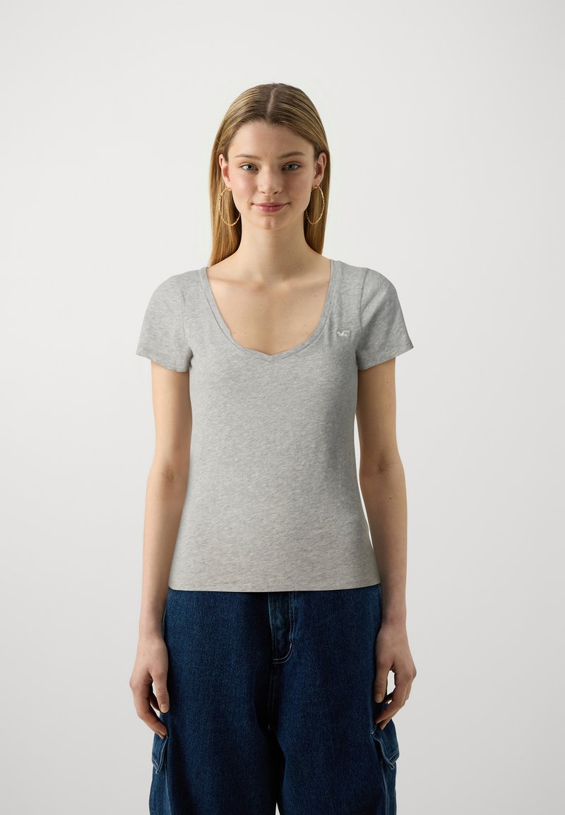 Szara krótkozasięgowa koszulka z dekoltem w serek, wykonana z miękkiego materiału. Posiada małe logo po lewej stronie na piersi. W połączeniu z ciemnoniebieskimi luźnymi dżinsami.