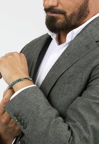 Grå texturerad kavaj med en vit skjorta under, prydd med ett grönt och silverfärgat armband på handleden som framhäver länkar och en jämn finish.