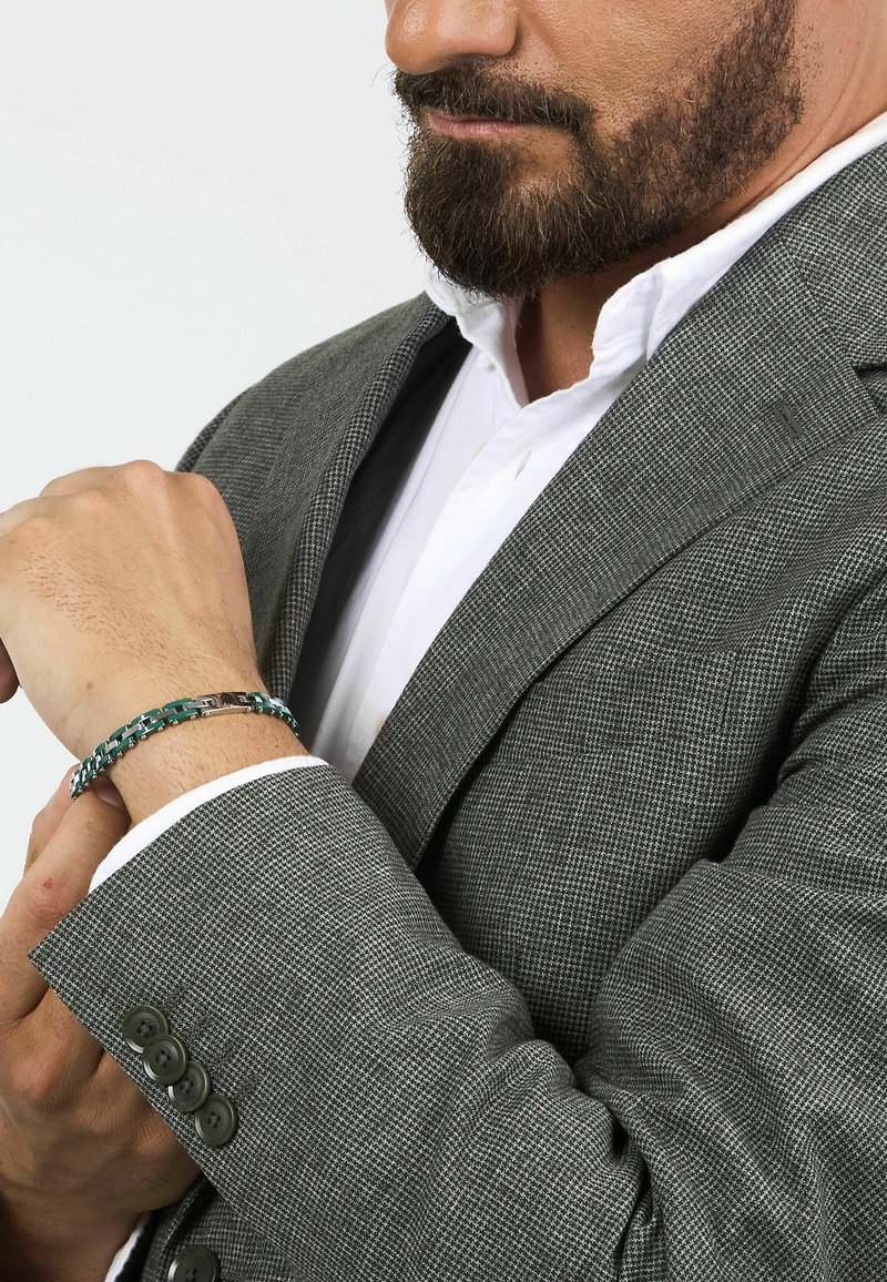 Grå texturerad kavaj med en vit skjorta under, prydd med ett grönt och silverfärgat armband på handleden som framhäver länkar och en jämn finish.