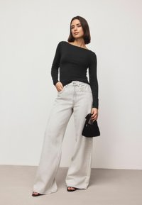 Sort langærmet top parret med lysegrå løse bukser med dekorativ sidepiping, mens der holdes en lille sort håndtaske.