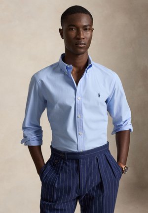 Homme portant une chemise bleu clair boutonnée avec les manches retroussées et un pantalon bleu marine à fines rayures, debout les mains dans les poches.