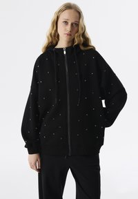Felpa nera con zip realizzata in materiale morbido, caratterizzata da decorazioni con borchie, vestibilità ampia, cappuccio con coulisse e polsini e orlo a costine.