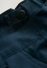 Nahaufnahme eines marineblauen Stoffes mit einer strukturierten Webart, einem schwarzen Knopf mit dem eingeprägten Namen "Clarks" und kontrastierendem Stich entlang der Kanten.