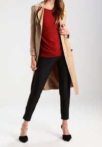 Trench-coat beige porté sur un haut rouge texturé, assorti à un pantalon noir à fines rayures et à des escarpins noirs à bouts pointus. Accessoires simples et lignes épurées.
