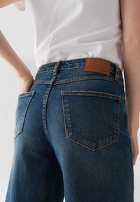 Close-up van een persoon die blauwe spijkerbroek en een wit shirt draagt, met een hand in de achterzak, leer label op de tailleband.