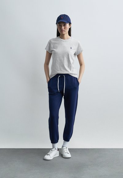 Νεαρή γυναίκα στέκεται με τα χέρια στις τσέπες, φορώντας γκρι μπλουζάκι, σκούρο μπλε παντελόνι φόρμας, λευκά αθλητικά παπούτσια, λευκές κάλτσες και σκούφο μπέιζμπολ ναυτικού χρώματος μπροστά σε απλό τοίχο.