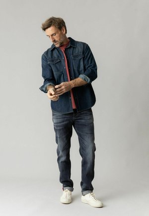 Mand iført en denimjakke og jeans, ruller ærmerne op, stående mod en ensfarvet lysegrå baggrund.