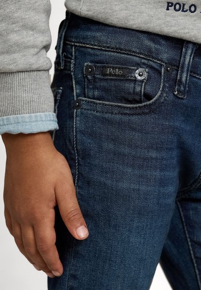 Jeans de un azul oscuro con un corte ajustado, que presentan una etiqueta negra de "Polo", dos bolsillos frontales y un detalle en puño de camisa en gris contrastante.