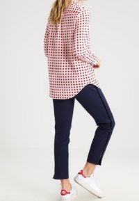 Nainen seisoo oikea jalka koukistettuna, päällä valkoisen ja punaisen ruudullinen paita, tummansiniset housut, joissa on sivuhalkio, ja valkoiset lenkkarit punaisilla yksityiskohdilla.