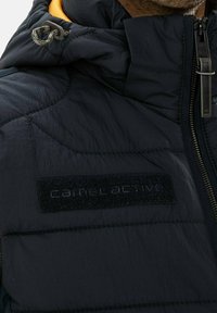 Tmavě modrá péřová bunda s texturovaným, prošívaným designem a zipovým zapínáním. Obsahuje černou látkovou nášivku s logem a kapuci se šňůrkou.