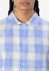 Short-sleeve plaid shirt in light blue and white, featuring a button-down collar, single chest pocket, and subtle purple grid accents.