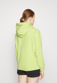 Grønn hettegenser med romslig hette, teksturert stoff og ribbede mansjetter. Båret med svarte shorts, som viser bakfra. Enkel design.