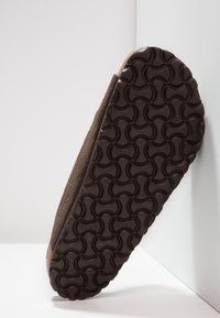 Chausson en Daim marron avec une semelle en caoutchouc noir texturé présentant des motifs circulaires. La semelle est légèrement surélevée au niveau du talon.