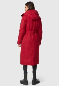 Manteau long rembourré rouge avec une capuche, taille ceinturée et fentes latérales. Tissu texturé et accent de marque sur la manches. Leggings noirs et bottes.