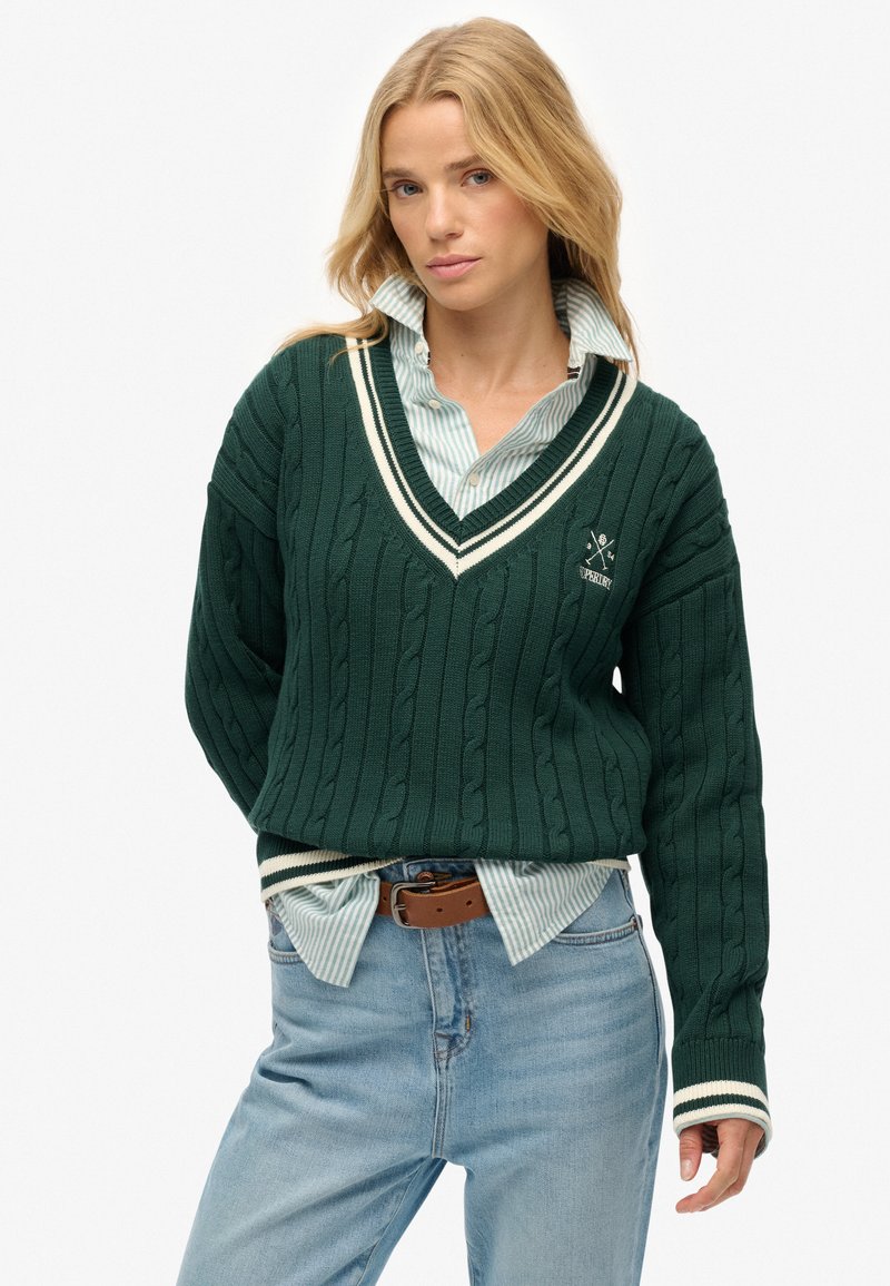 Groene gebreide trui met kabelstructuur, witte afwerking op de V-hals en manchetten, over een blauw-witte gestreepte overhemd, gecombineerd met jeans.