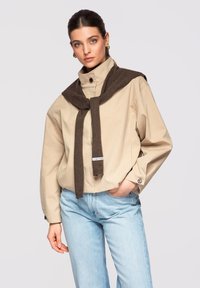 Femme aux cheveux foncés attachés en arrière, portant une veste beige boutonnée, un jean bleu clair et un pull marron posé sur les épaules, debout avec la main dans la poche.