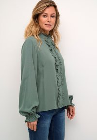 Green blouse made of lightweight fabric, featuring a ruffled front, high collar, and puffed sleeves with elastic cuffs. Worn with blue jeans.