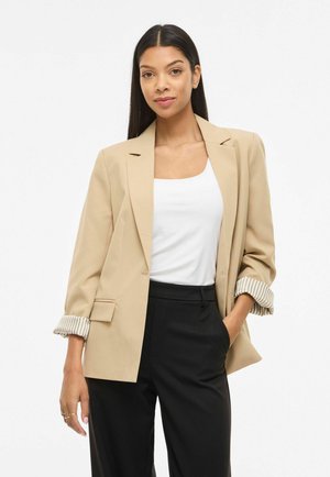 Kvinne med langt mørkt hår, iført en beige blazer med oppbrettede stripete mansjetter, hvit topp og svarte bukser, stående med den ene hånden i lommen.