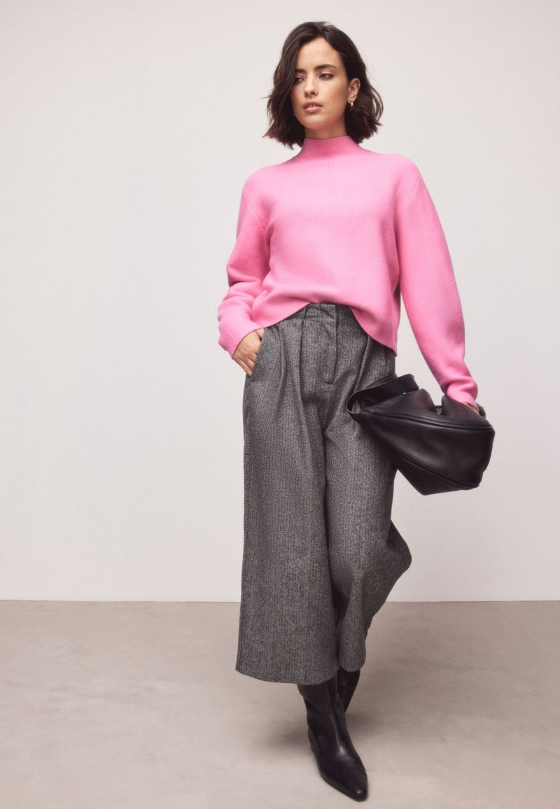 Nő élénk rózsaszín pulóverben, szürke bőszárú nadrágban, fekete csizmában, fekete bőrtáskát tartva egyszínű világos háttér előtt.