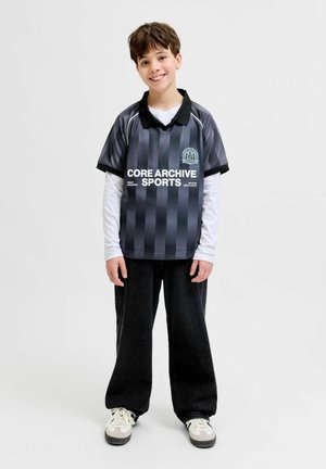 Garçon souriant debout, portant un polo de sport rayé noir et gris sur une longue chemise blanche, un pantalon noir ample et des baskets blanches sur un fond uni.