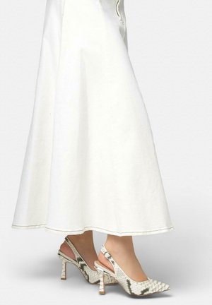 Persona con falda midi blanca y zapatos de tacón slingback de punta puntiaguda con estampado de piel de serpiente sobre fondo gris.