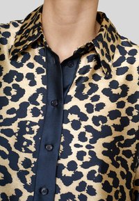 Kvinde iført en beige og sort leopardmønstret satinv blouse med sort knaplukning og spidse krave, nærbillede af overkroppen.