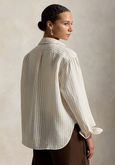 Mujer de cabello oscuro recogido en un moño, vestida con una camisa de manga larga de rayas verticales beige y blancas y pantalones marrones, de perfil sobre un fondo neutro.